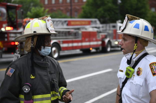 Zwei Feuerwehrleute in Helmen, einer mit Maske, unterhalten sich auf einer Straße mit Fahrzeugen, Bäumen, Gebäuden und einem Pfahl im Hintergrund.