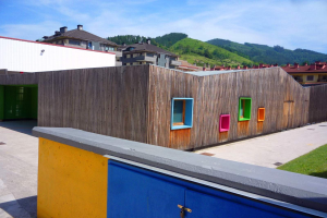 Ein hölzernes Gebäude mit bunten Fenstern, einer blauen Tür, Gras, einer Wand, Bäumen und einem bewölkten Himmel, Teil des Zell am See Kindergartens