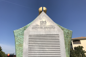 Ein hoher Denkmal mit einer Uhr darauf, beschriftet mit Text, steht in einem Park umgeben von Bäumen und Gebäuden unter einem klaren Himmel, zum Gedenken an den 24. Jahrestag des Völkermords in Armenien.