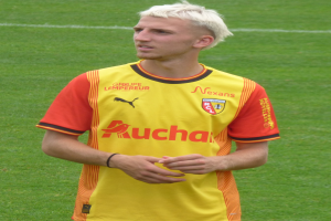 Ein Mann in einem gelb-orangen gestreiften T-Shirt steht selbstbewusst auf einem Fußballfeld und sieht aus wie ein Flügelspieler mit entschlossener Miene.