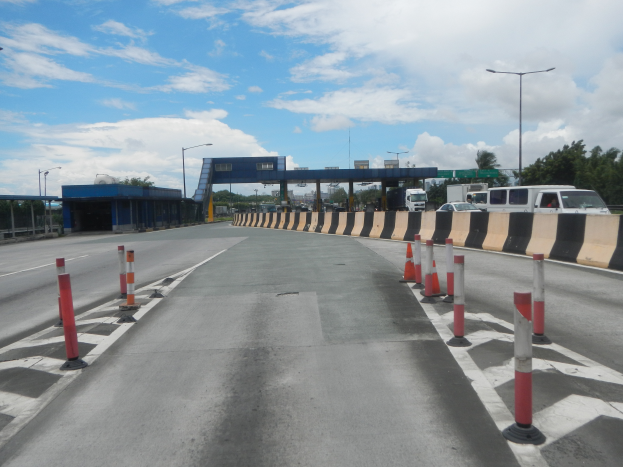 Eine Straße mit Verkehrsleitkegeln am Rand, eine Mittelleitplanke, Fahrzeuge, Laternenpfähle, Bäume, eine Brücke und ein bewölkter Himmel im Hintergrund.