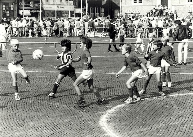 Schwarzes Bild von jungen Jungs, die Fussball auf der Straße spielen, umgeben von einer Zuschauermenge hinter einem Geländer, Gebäuden, Pfählen und Texttafeln im Hintergrund.
