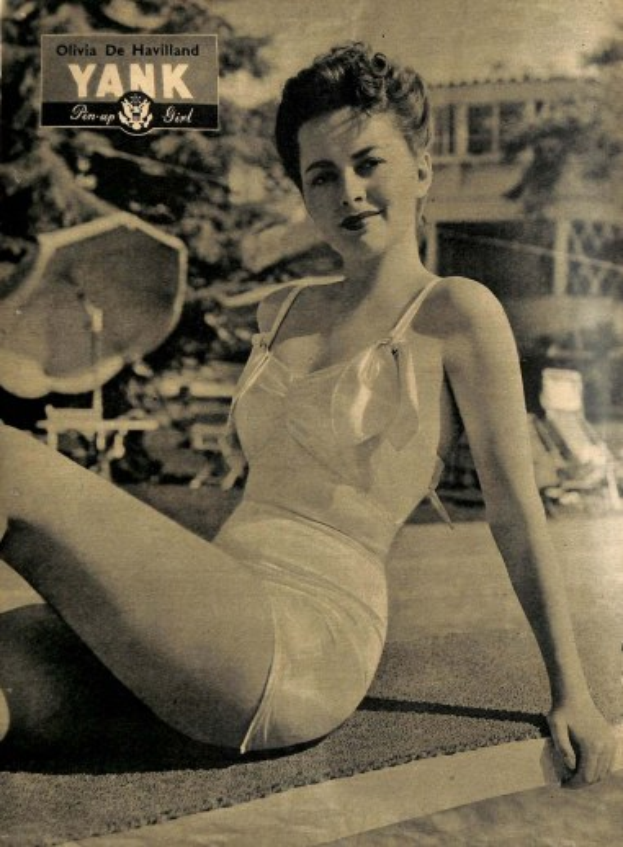 Olivia de Havilland in einem Badeanzug sitzend auf dem Boden und lachend, mit Bäumen, Stühlen, einem Gebäude und Text im Hintergrund.