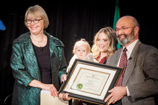 Zwei Personen, ein Mann mit einem Zertifikat und eine Frau mit einem Gegenstand, stehen lachend nebeneinander auf einer Preisverleihung mit einer Flagge und einem schwarzen Vorhang im Hintergrund.