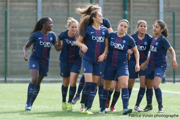 Gruppe junger Frauen, die auf einem Rasenfeld Fußball spielen, mit Maschendrahtzaun und einer Wand im Hintergrund, beschriftet mit "Paris Saint-Germain Frauenfußball" in der rechten unteren Ecke.