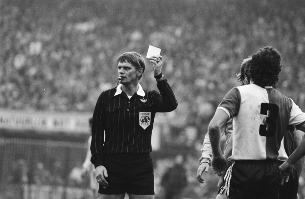 Schwarzes und weißes Bild eines Schiedsrichters mit roter Karte vor einer Menge in Fußballuniformen und einem Zaun im Hintergrund.