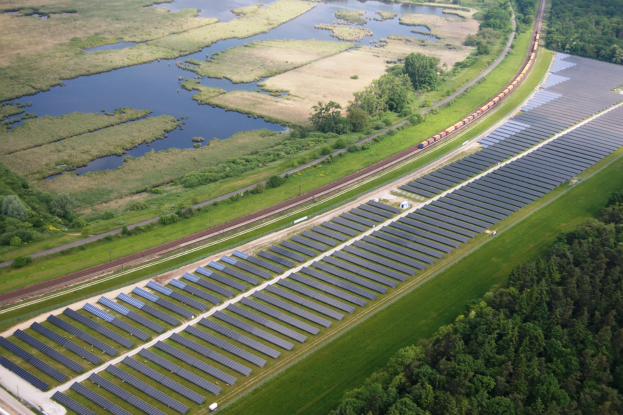 Eine Luftaufnahme eines Solarparks mit Panelen in einem Feld, umgeben von Bäumen, Gras und Wasser, mit einem Zug auf einer nahen Eisenbahnschiene.