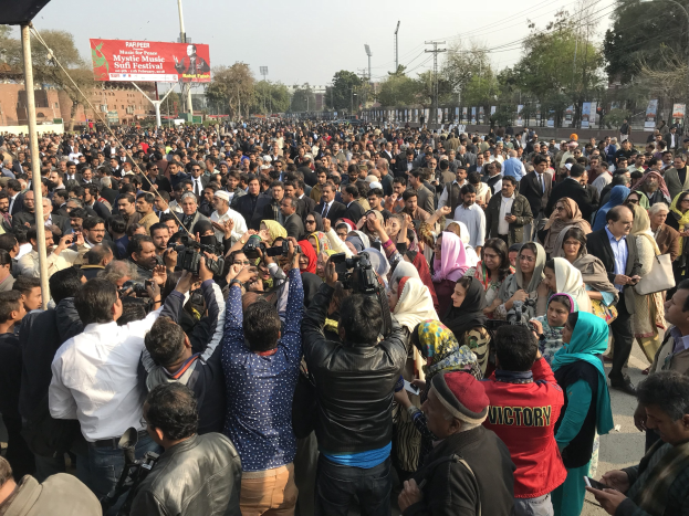 Eine große Menschenmenge steht vor einem Gebäude, einige halten Kameras und Handys, mit einem Banner, das "Kashmiri muslims protest in Lahore" zeigt, und anderen städtischen Elementen im Hintergrund.