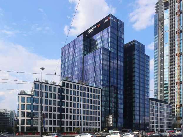 Außenansicht des Deutschen Bank-Hauptsitzes in Frankfurt, Deutschland, mit einem mehrstöckigen Gebäude mit zahlreichen Fenstern, umgeben von städtischer Infrastruktur wie Straßenlaternen, Fahrzeugen, Bäumen und einem bewölkten Himmel.
