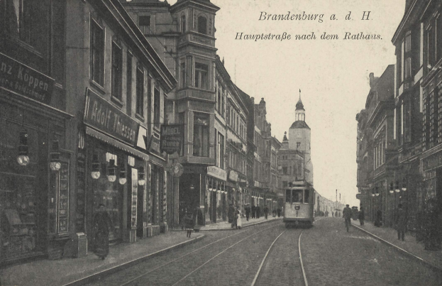 Schwarzes und weißes Foto einer Stadtstraße in Brandenburg, Deutschland mit einer Straßenbahn, Passanten, Gebäuden, Schildern, Straßenlaternen und einem bewölkten Himmel.