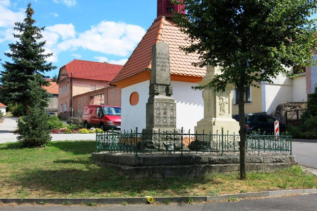 Ein kleiner Stadtplatz mit einem Holocaust-Mahnmal in der Mitte, umgeben von einem Metallzaun, Gras, Bäumen, Fahrzeugen, einem Schild, Häusern und einem bewölkten Himmel.