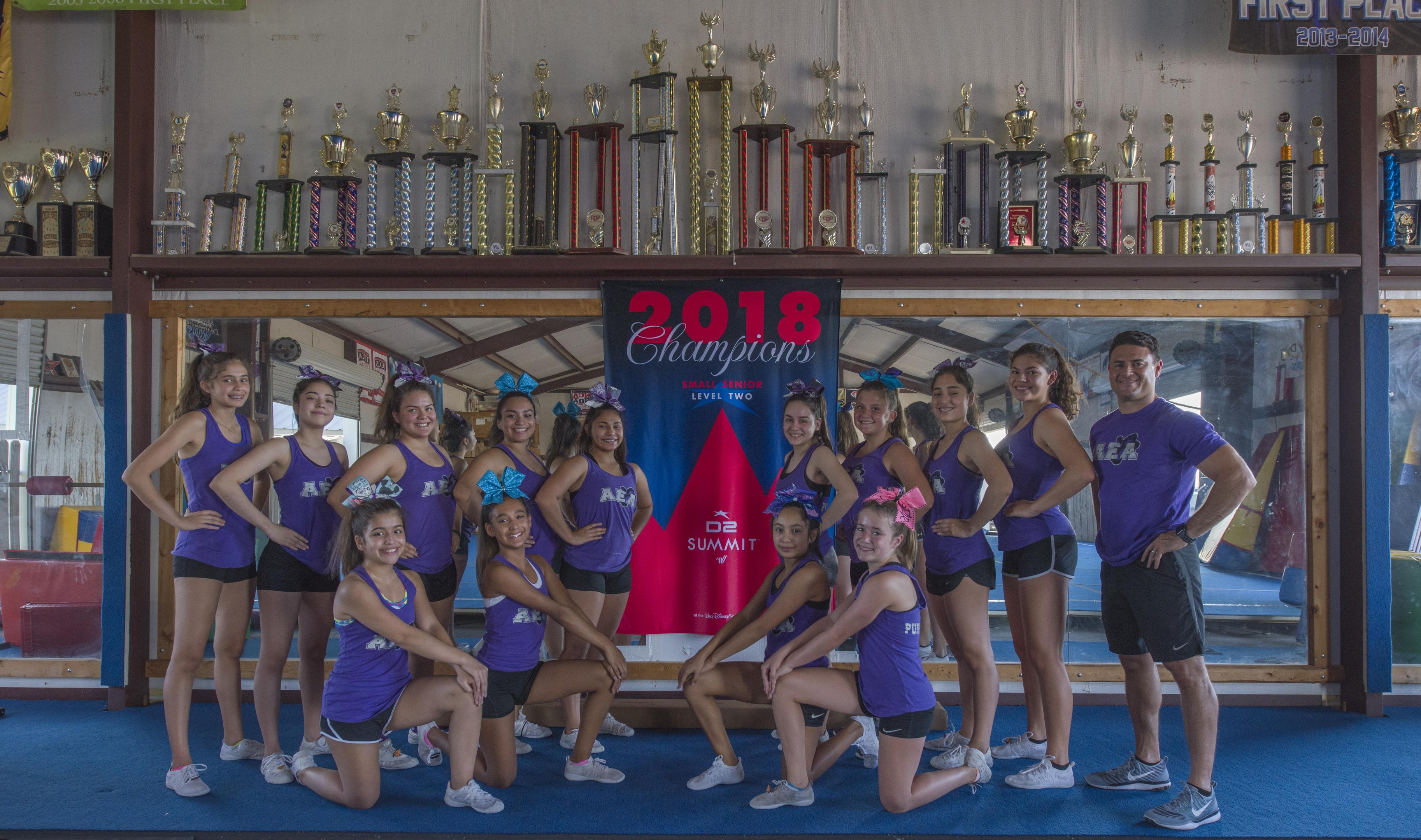 Gruppe von Cheerleadern in lila T-Shirts und schwarzen Shorts, die vor einem Vitrinenschrank mit einer '2018 Summit Cheerleading Champions'-Banner im Hintergrund posen.