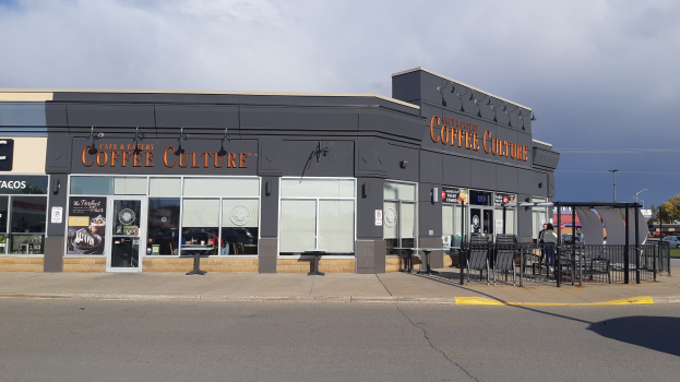 Ein Kaffeehaus an einer Straßenecke mit einer Straße im Vordergrund, einem Gebäude im Hintergrund, Fahrzeugen, Bäumen und einem blauen und weißen Himmel.