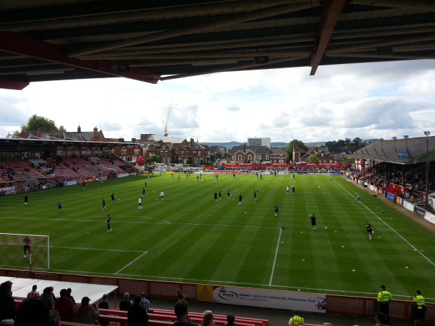 Ein Fußballspiel wird im St Mary's Stadium gespielt, mit Zuschauern auf Bänken und Stehplätzen und einem Torpfosten links sowie Gebäuden, Bäumen und einem bewölkten Himmel im Hintergrund.