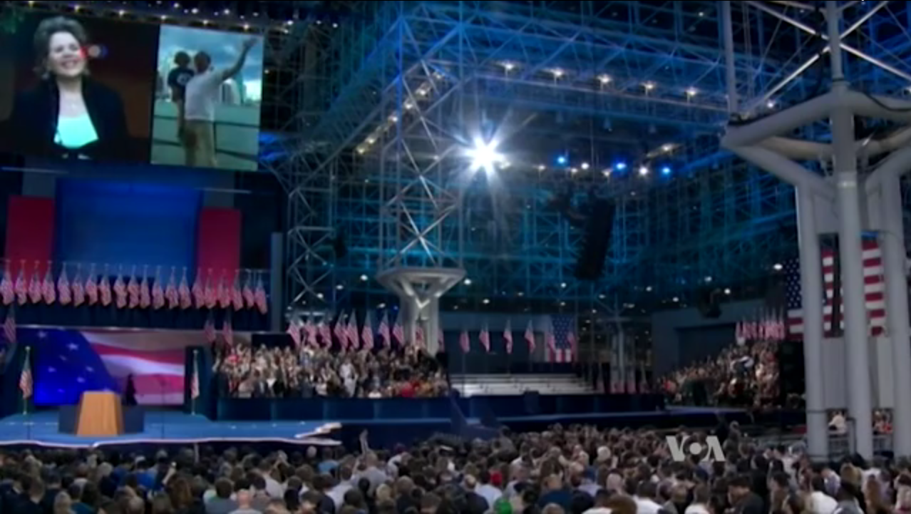 Eine große Menschenmenge vor einer Bühne mit einem Rednerpult, Mikrofon, Flaggen, einem Bildschirm und Beleuchtungsausrüstung, die auf eine Präsidentenrede warten.