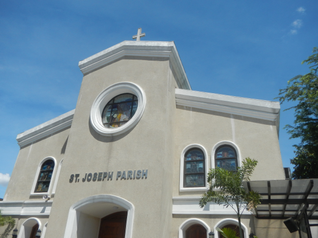 St. Joseph Parish Church in Key West, Florida, a building with windows, doors, and a cross on top, surrounded by trees and plants, with a sky filled with white, fluffy clouds in the background.