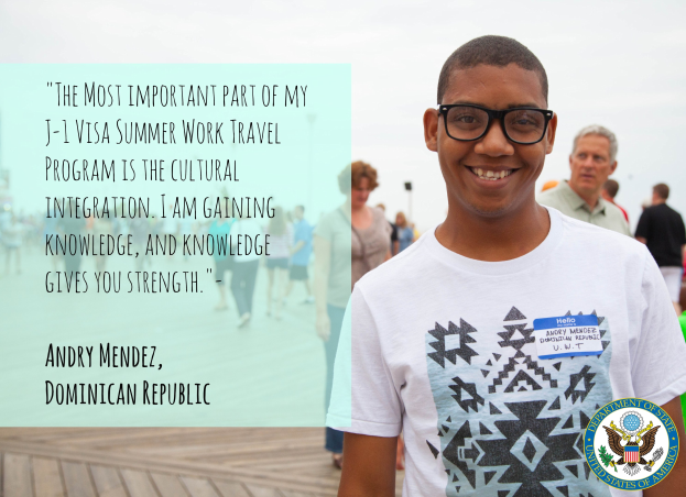 Eine Person in einem weißen T-Shirt lächelt, während sie mit einer Gruppe auf einer hölzernen Oberfläche geht, mit einem Zitat von Andry Mendez über kulturelle Integration im J-1-Visa-Programm auf der linken Seite, vor Buildings und einem klaren blauen Himmel.