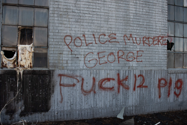 Graffiti an einer Hauswand mit der Aufschrift "Polizei ermordet George Fuck 12 P19" mit Fenstern und verstreuten Gegenständen davor.