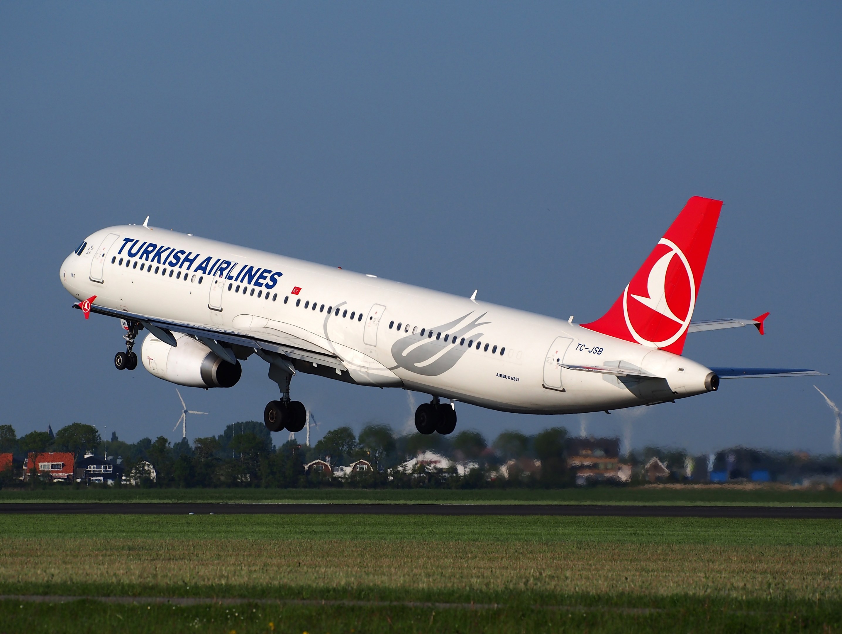 Turkish Airlines Airbus A330-300 in Startposition am Frankfurter Flughafen, mit Gras darunter, Gebäuden und Bäumen im Hintergrund, Windrädern in der Ferne und einem klaren blauen Himmel.