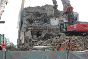 Eine Abbruchstelle mit einem Kran im Vordergrund, umgeben von anderen Kränen, Fahrzeugen, Schutt und einem Banner, mit Gebäuden, Bäumen und Himmel im Hintergrund.