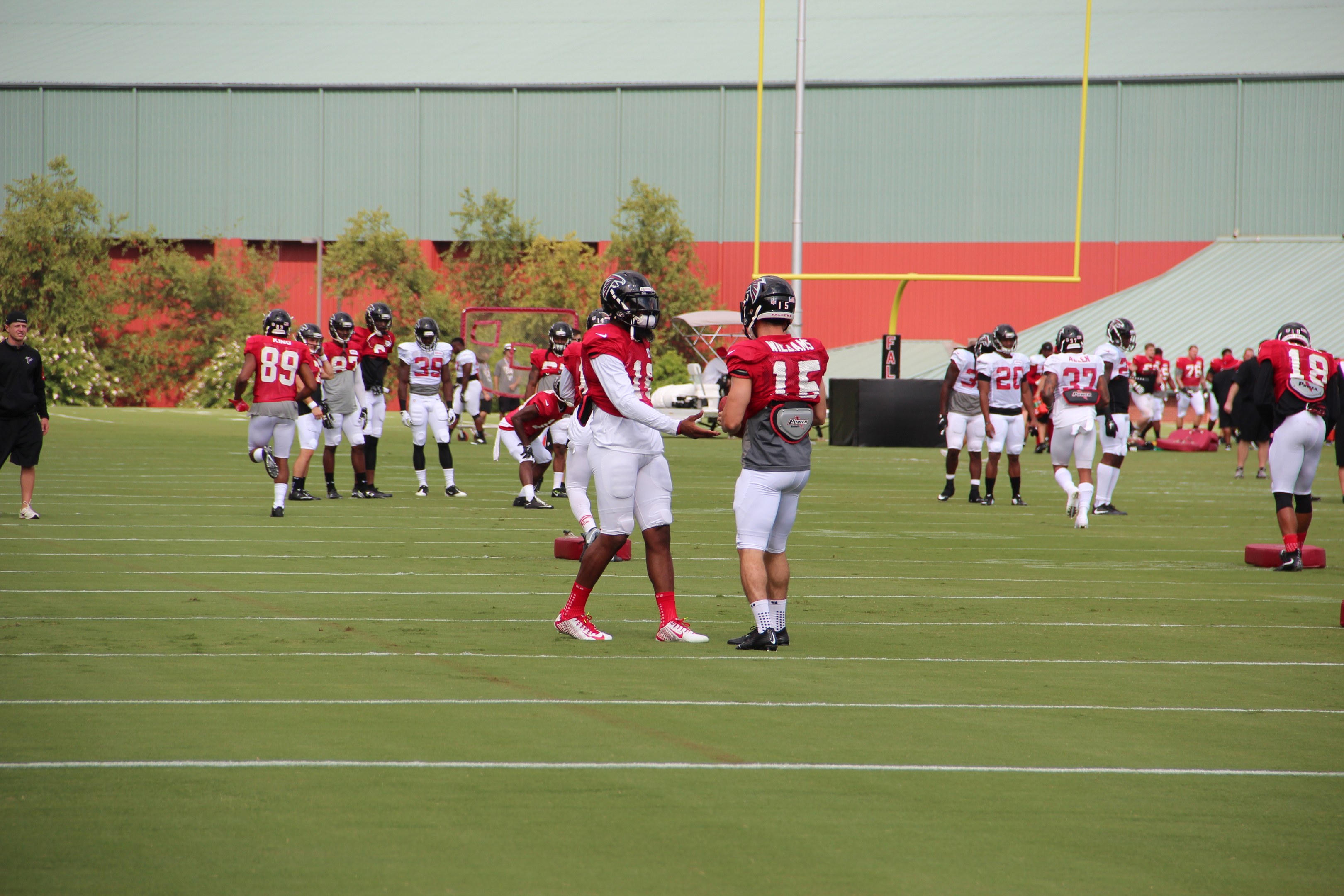 Gruppe von Menschen, die Fußball auf einem Rasenfeld mit Helmen und Fußballschuhen spielen, umgeben von Bäumen, Pfählen, einer Wand und einem klaren blauen Himmel.