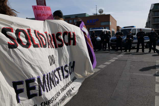 Gruppe von Menschen marschiert auf einer Straße und hält eine 'Solidarität und Feminismus'-Tafel, mit parkenden Fahrzeugen, Gebäuden, einer Schüsselantenne und einem klaren blauen Himmel im Hintergrund.