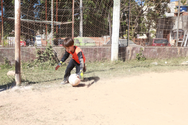 Junger Junge spielt Fußball auf einem Schotterfeld mit umgebenem Gras, Pflanzen, Pfählen, einem Zaun, einer Wand, Bäumen, Fahrzeugen, Gebäuden und einem bewölkten Himmel, trägt Schuhe und ist aktiv im Spiel.