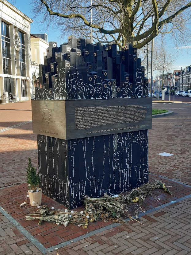 Denkmal für Holocaust-Opfer in einem Stadtplatz, mit einem Baum davor, einem Topf mit einer Pflanze, verstreuten trockenen Blättern und Gebäuden im Hintergrund.