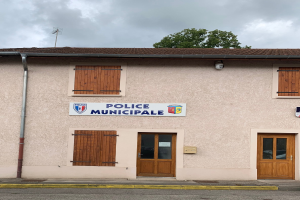 Gebäude der Police municipale mit einem Schild, Fenstern, Türen, einer Namensplakette, einem Rohr, einem Baum und einem bewölkten Himmel im Hintergrund.
