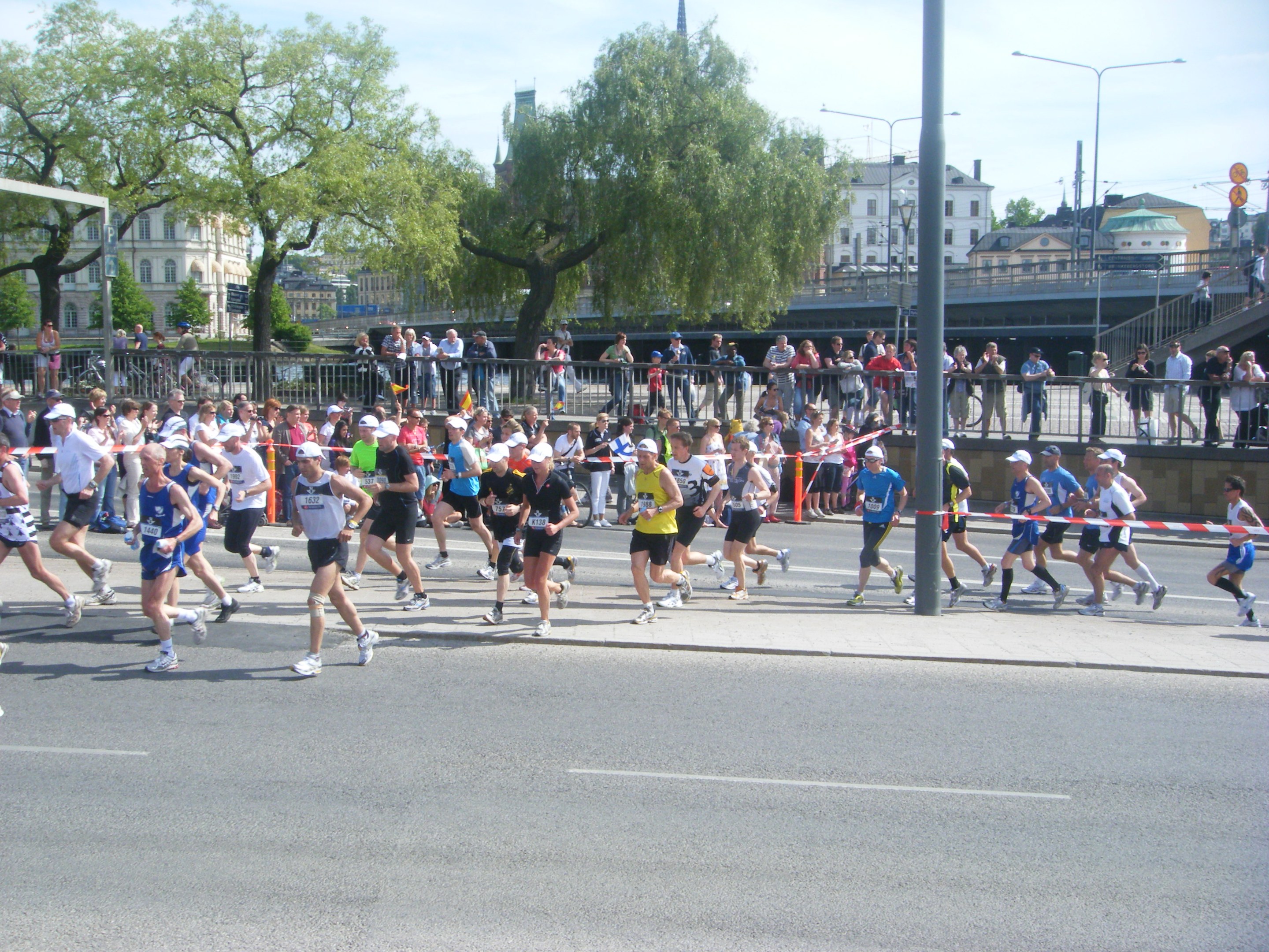 Gruppe von Menschen beim Laufen eines Marathons auf einer Straße mit einer Ziellinienbanderole, umgeben von Absperrungen und Barrieren, unter einem bewölkten Himmel.