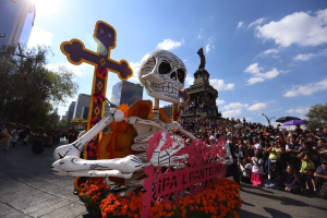 Ein Tag der Toten-Umzug in Mexiko-Stadt mit einer zentralen Skelettstatue, die mit Blumen und einem Kreuz geschmückt ist und von Zuschauern mit Schirmen und Passanten an der Straße flankiert wird, vor einem Hintergrund aus Bäumen, Gebäuden und einem bewölkten Himmel.