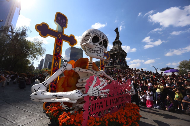 Ein Tag der Toten-Umzug in Mexiko-Stadt mit einer zentralen Skelettstatue, die mit Blumen und einem Kreuz geschmückt ist und von Zuschauern mit Schirmen und Passanten an der Straße flankiert wird, vor einem Hintergrund aus Bäumen, Gebäuden und einem bewölkten Himmel.