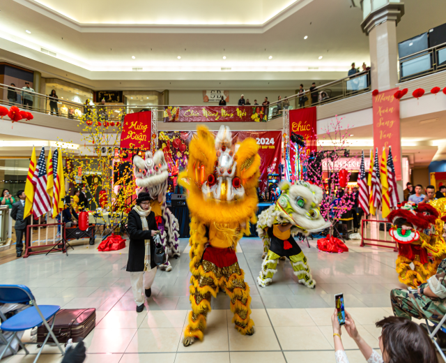 Ein lebendiges chinesisches Neujahrsfest in einem Einkaufszentrum mit Menschen in Kostümen, die tanzen, und anderen, die auf Stühlen sitzen und mobile Telefone halten, umgeben von Flaggen, Bannern, Dekorationen und Lichtern.