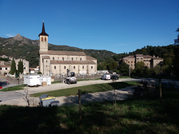 Kleiner Ort mit einer zentralen Kirche, umgeben von Gebäuden, Bäumen, Fahrzeugen und anderen Gegenständen, vor einem Hügel- und einem klaren blauen Himmel.