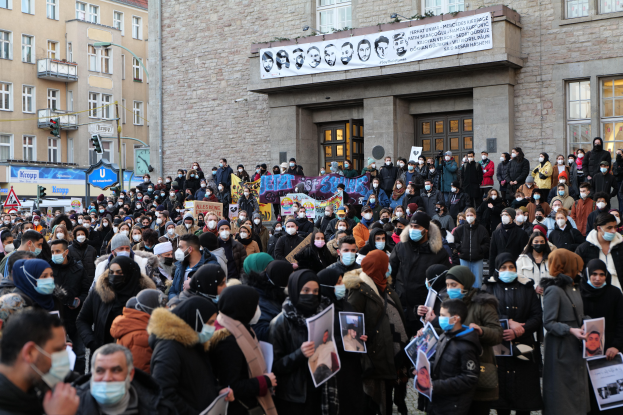Eine große Gruppe von Menschen mit Masken und Schildern versammelt sich vor einem Gebäude mit Fenstern, Türen, Bannern, Laternenmasten und Verkehrsschildern und protestiert gegen eine Regierungsentscheidung, das Tragen von Masken zu verbieten.