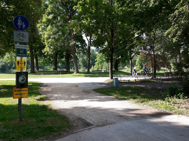 Ein Park mit einem asphaltierten Mittelweg, der von Bäumen und Gras gesäumt ist und auf der linken Seite eine Tafel an einem Pfahl sowie im Hintergrund Radfahrer zeigt.
