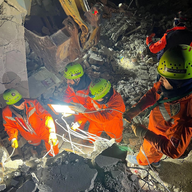 Gruppe von Menschen in orangefarbener Uniform und grüner Helme bei der Arbeit an Schutt mit einem Kran im Hintergrund bei einer Rettungsaktion nach einem Einsturz.