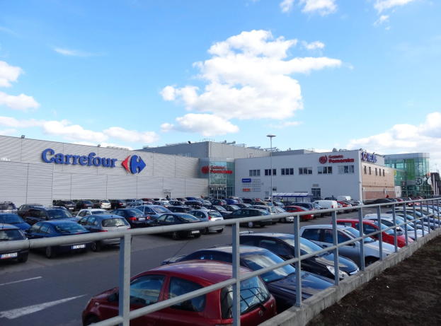 Großes Autohaus mit zahlreichen Fahrzeugen vor der Tür, umgeben von städtischen Gebäuden, Straßeninfrastruktur und Fahrzeugen auf der Straße unter einem bewölkten Himmel.