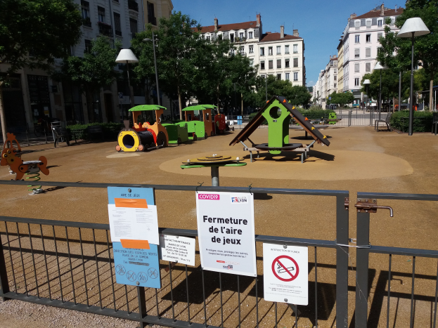 Ein Spielplatz auf einem Stadtplatz, umgeben von einem Zaun mit mehreren Schildern, mit Bäumen, Laternenmasten, Gebäuden und verstreuten Gegenständen im Hintergrund unter einem sichtbaren Himmel.
