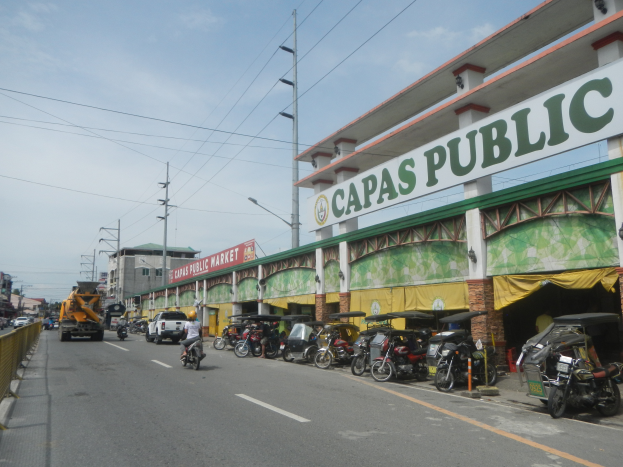 Eine belebte Stadtstraße mit Fahrzeugen, einem Geländer auf der linken Seite, Gebäuden, Pfosten, Laternen, Schildern und einigen Menschen unter einem bewölkten Himmel, mit einem Gebäude mit der Aufschrift "Capas Public".
