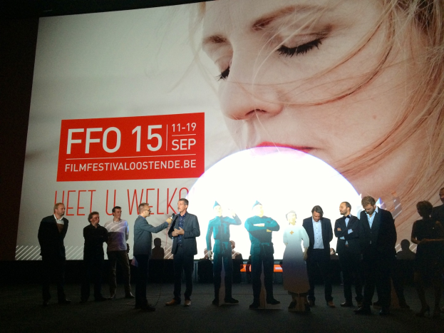 Eine Gruppe von Menschen auf einer Bühne mit einem Mann, der ein Mikrofon hält, im Zentrum, ein Display hinter ihnen zeigt den Text "Foto Filmfestivaloostende 2015" und ein Bild einer Frau.