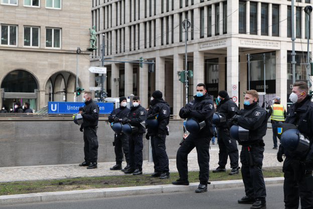Eine Gruppe von Polizisten in schwarzen Uniformen und Masken steht vor einem Gebäude mit Glasfenstern und Säulen in Berlin, Deutschland, einige halten Helme, mit Laternen, Verkehrszeichen, Texttafeln, einer Statue, anderen Menschen und einem grasbewachsenen Boden im Hintergrund.