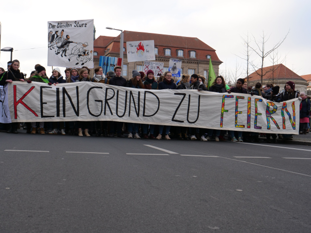 Demonstranten mit einem Transparent "Kein Grund zu Feiern" gegen deutsche Sparmaßnahmen, mit Straßeninfrastruktur und Gebäuden im Hintergrund vor einem klaren Himmel.