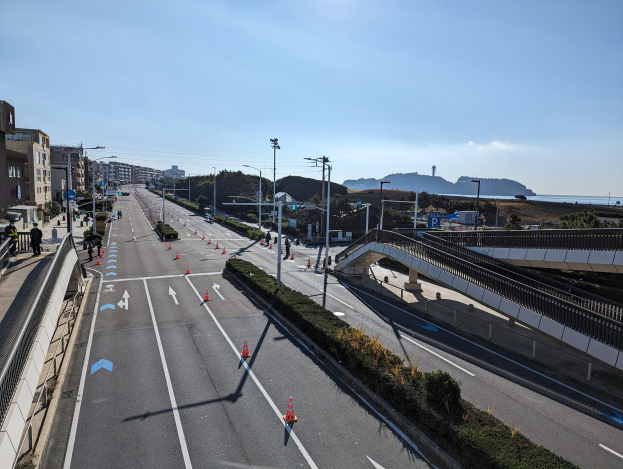 Straße mit Verkehrskegeln, eine Brücke mit Geländern, Laternen, Gebäude mit Fenstern und einige Menschen; Hügel und bewölkter Himmel im Hintergrund, Teil des Takayama-Brücken-Projekts.