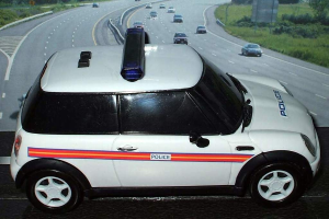 Weißes Polizeiauto mit "Polizei" Aufschrift auf einer Straße außerhalb einer Stadt, umgeben von fließendem Verkehr, Gras und Bäumen.