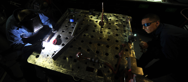 Zwei Männer in Schutzausrüstung arbeiten an einem Metallstück in einer dämmrig beleuchteten Werkstatt, mit verschiedenen Werkzeugen und Materialien auf dem Tisch davor.