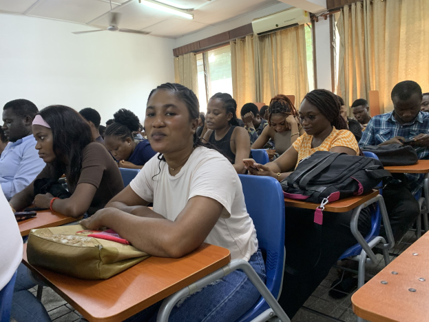 Eine Gruppe von Studenten sitzt an Tischen in einem classroom an der Universität von Ghana, mit Taschen und Gegenständen auf den Tischen, Fenstern mit Vorhängen im Hintergrund und an der Decke montierten Lampen, Ventilatoren und einer Klimaanlage.