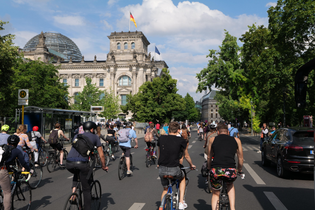 Gruppe von Menschen, die Fahrräder auf einer von Bäumen gesäumten Straße in Berlin, Deutschland, fahren, mit Gebäuden und einer Bushaltestelle, unter einem bewölkten Himmel und einer Flagge an einem Gebäude.