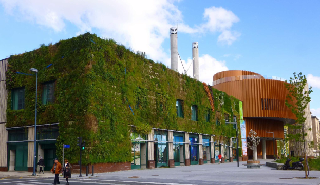Ein großes Gebäude mit einer grünen Wand an seiner Seite, umgeben von Straßeninfrastruktur, Fahrzeugen, Fußgängern, Bäumen und einem bewölkten Himmel, als Teil des Green Building Project.