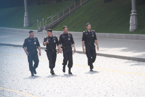 Drei Polizisten in Uniform und Mützen, die eine Straße mit Pfosten, Geländern, Treppen und Gras im Hintergrund entlanggehen.
